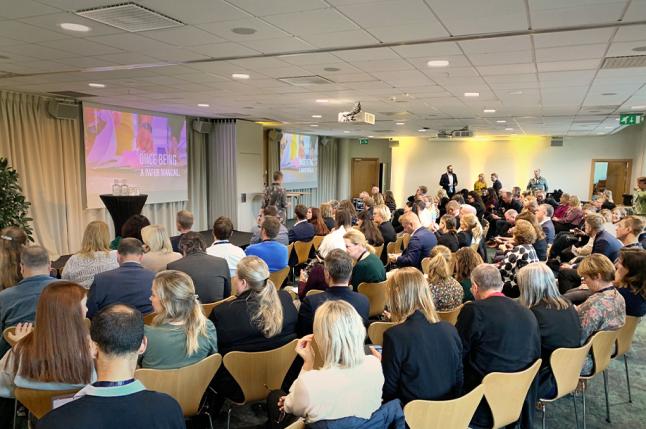 Ett konferensrum med många sittande människor. Alla vänder ryggen mot kameran.